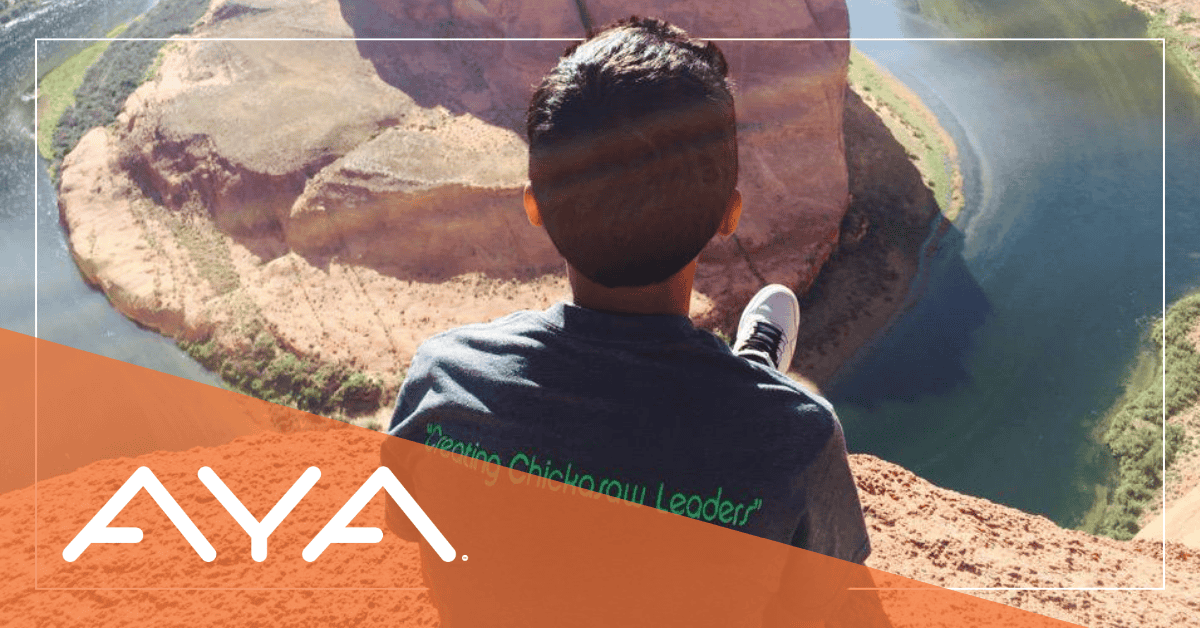 Person wearing'Creating Chickasaw Leaders' t-shirt sits overlooking a dramatic red rock canyon with blue walk below. View is from behind, showing AYA logo with orange overlay. Image illustrates connection between nature walks and mindfulness.