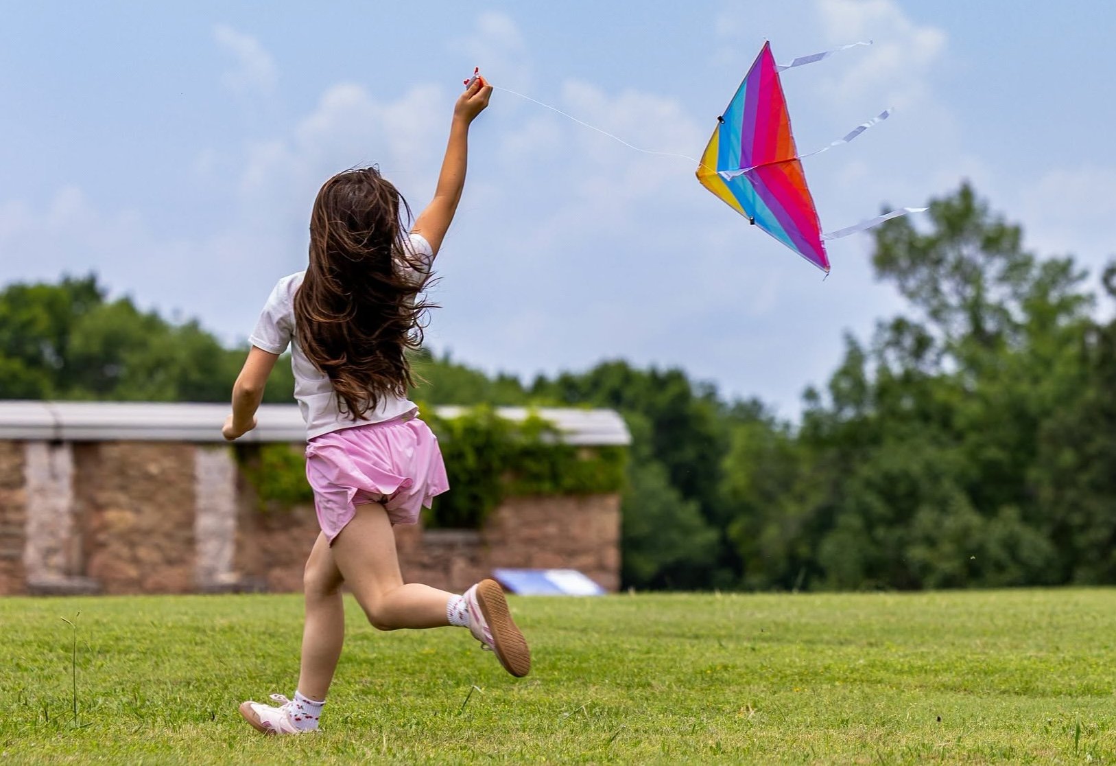 A young girl with brown hair running in a field of green grass with a rainbow kite on a string flying behind her.