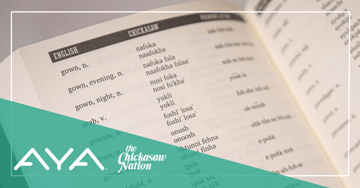 A English to Chickasaw dictionary is open to a page providing Chickasaw language translations of English words with a pronunciation guide. Diagonally across the bottom left corner is a green banner with the AYA and Chickasaw Nation logos.