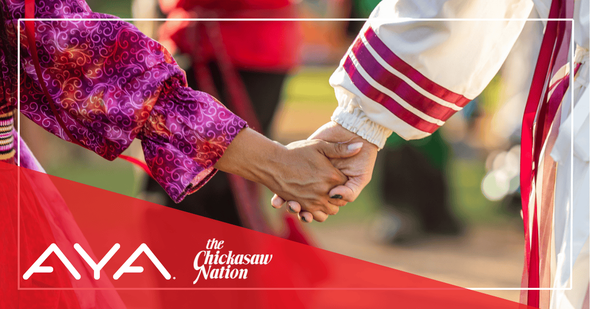 Two people, one in a traditional Chickasaw ribbon shirt with red ribbons and one in a pink and purple traditional Chickasaw dress are holding hands. In the left corner of the image is a red triangle with the AYA and Chickasaw Nation logos.