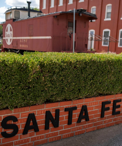 A red brick wall with 'SANTA FE' lettering below a neatly trimmed hedge, with a vintage red train car visible in the background.