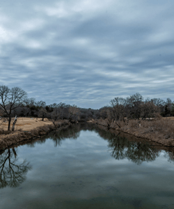 A calm river reflects the Penning Creek with bare winter trees and moody gray clouds, creating a mirror-like symmetry in the landscape.