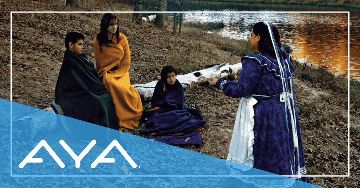 A Chickasaw storyteller in traditional dress tells a story to three people sitting on the ground
