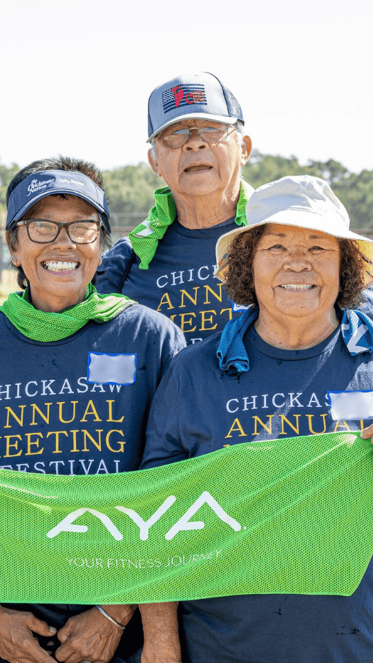Three Chickasaw citizens wearing Annual Meeting and Festival t-shirts and caps, holding a bright green AYA banner while smiling at the camera.