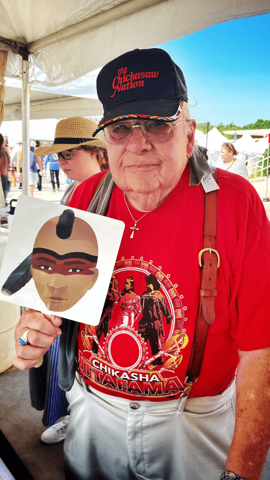 Elder Chickasaw citizen wearing a Chickasaw Nation cap and red Chikasha Inkana t-shirt, holding up a fan with the face of Hikatubby, AYA Walking Partner.
