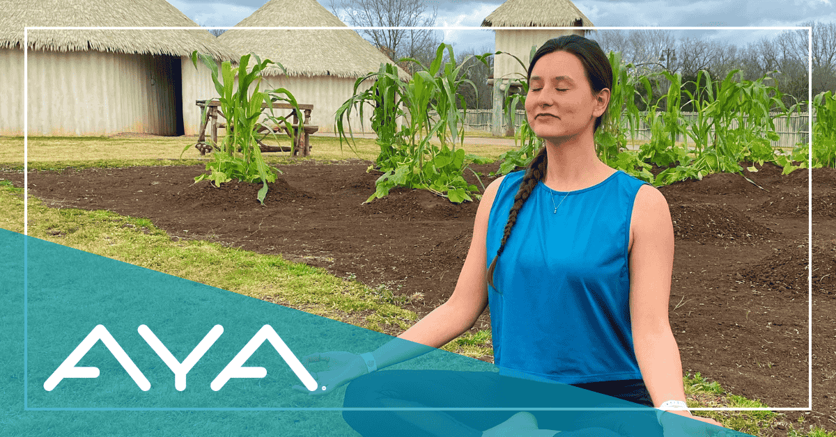 A women sits on the ground outside in a yoga pose with her eyes closed.
