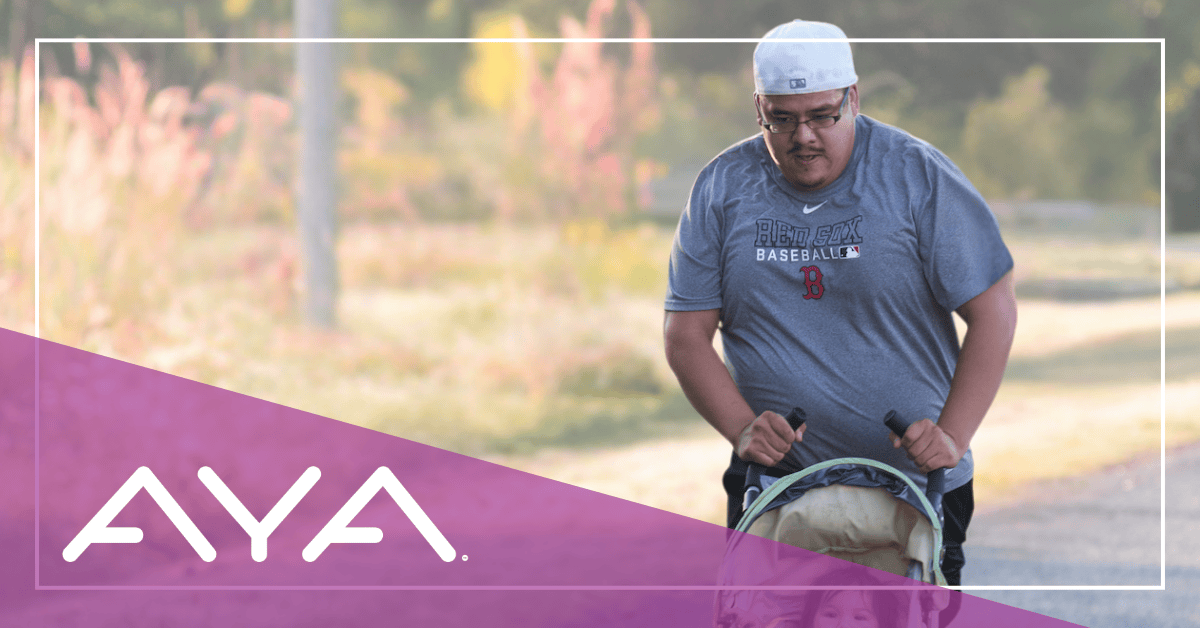A man in a gray t-shirt walks outside near some trees while pushing a baby stroller