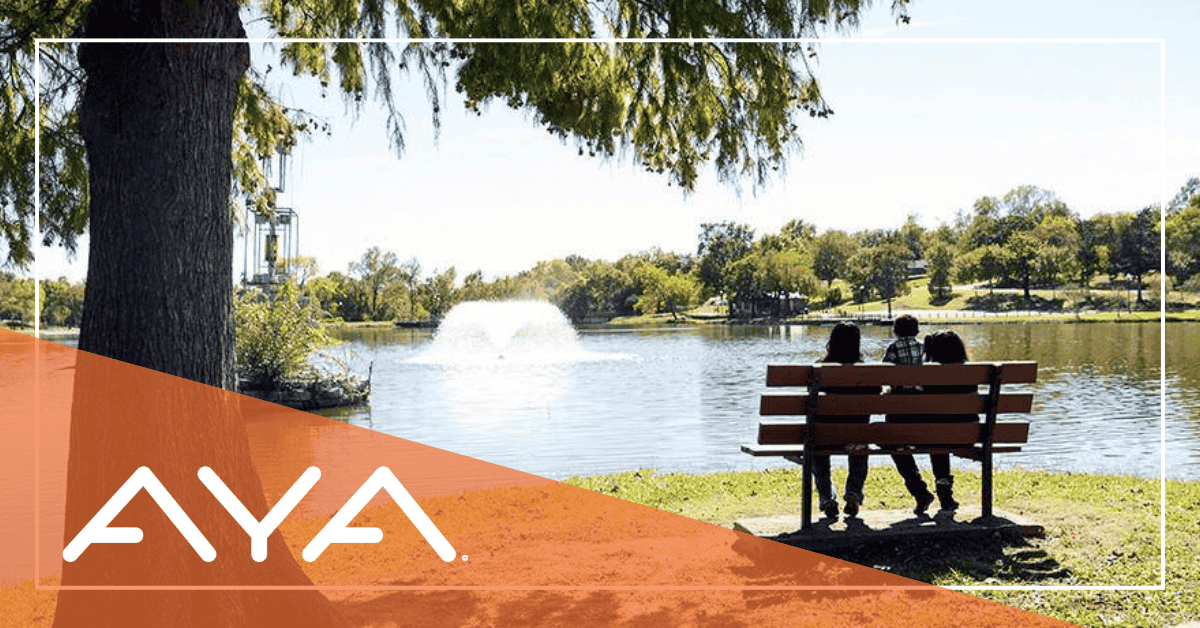 Two people sit on a bench by a lake with a fountain and trees.
