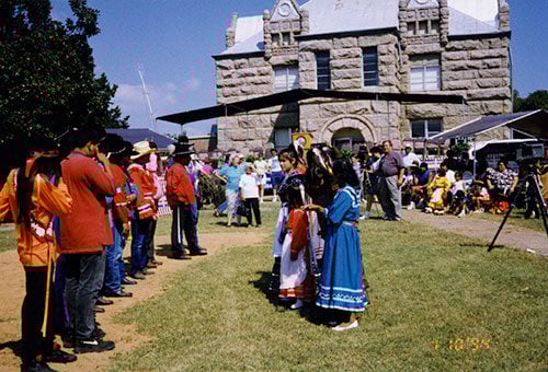 Color photograph showing a gathering at the historic Chickasaw Capitol Building in Tishomingo, Oklahoma. People in traditional dress, including bright red and blue garments, stand on the lawn in front of the distinctive stone building's facade.