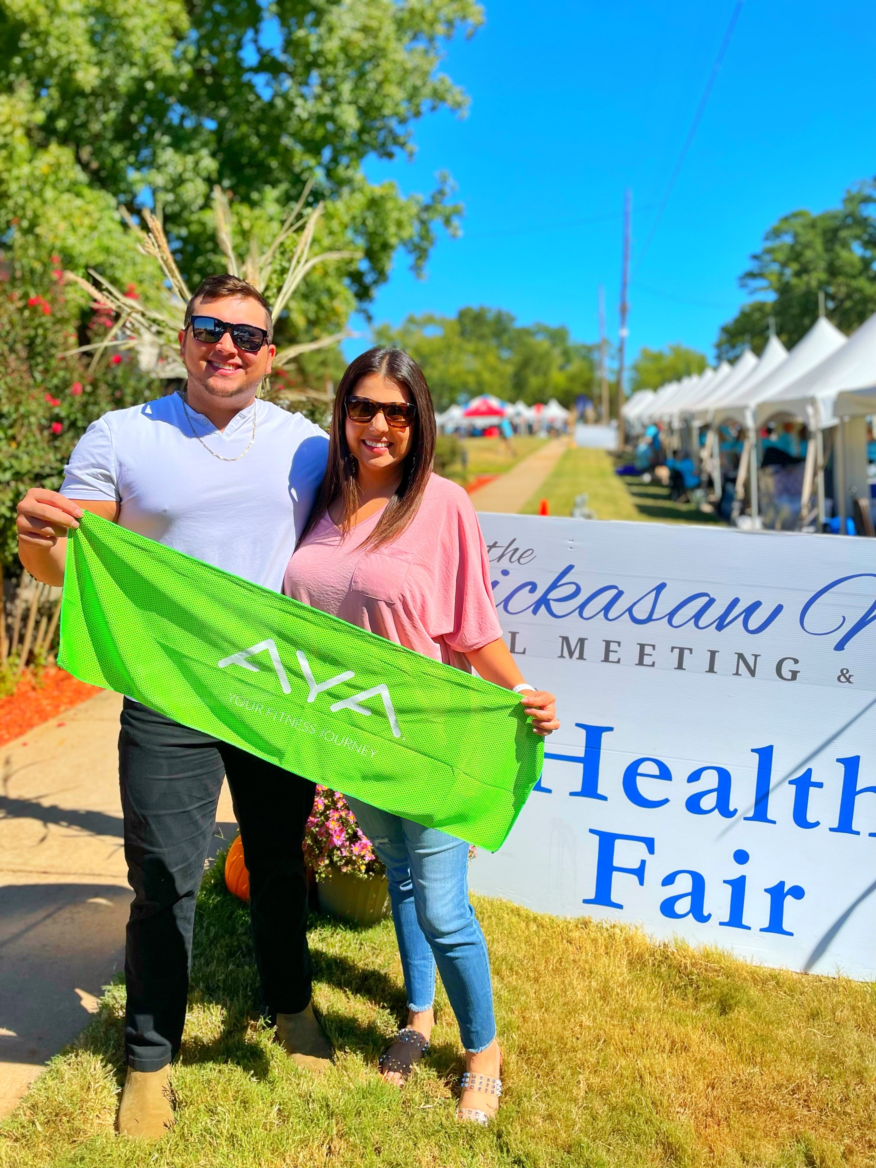 Two Chickasaw citizens holding a green AYA cooling towel in front of a Chickasaw Health Fair sign