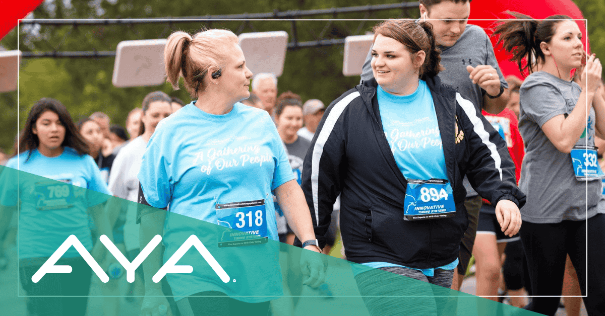 Two women wearing matching light blue t-shirts with race numbers participate in a walk event. They're smiling and walking together, with other participants visible in the background. The AYA logo appears in white against a teal overlay.