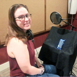 A Chickasaw woman voice actor for the AYA walking app smiles at the camera while seated at a professional recording station. She wears glasses and a burgundy top, and is positioned at a microphone with a pop filter and acoustic panels.