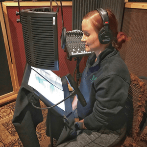 A Chickasaw woman in a recording booth sits with headphones, reading from a tablet in front of a microphone with an acoustic filter screen, capturing audio in a professional studio setting.