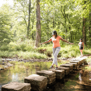 A person in orange athletic wear carefully crosses a stone stepping path over a stream, arms outstretched for balance. Another person visible in the background. Sunlit forest setting with green foliage