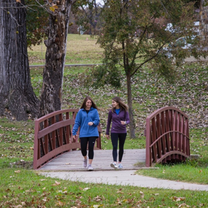 Two women walking and talking together on a wooden footbridge, wearing casual athletic wear in blue and purple. The bridge is surrounded by autumn trees and fallen leaves in a park-like setting