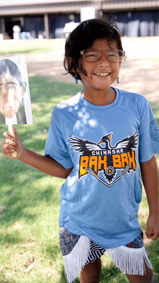 Young Chickasaw child wearing glasses and a light blue Chikasha Bak Bak t-shirt with white fringed shorts, smiling while holding a fan featuring the face of Eliza, AYA Walking Partner.