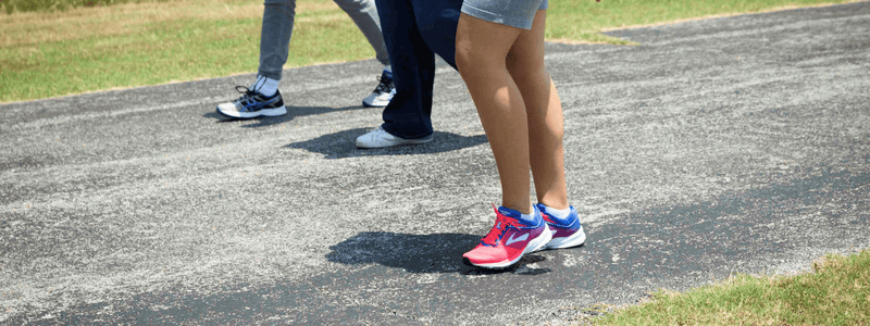 Close-up of three people's legs and feet on a walking path, focusing on bright pink and blue athletic shoes in the foreground, demonstrating proper walking footwear. Other walkers wear supportive sneakers in the background.