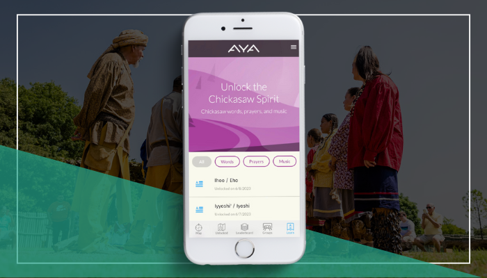 iPhone displaying the AYA app's 'Unlock the Chickasaw Spirit' screen with options for words, prayers, and music. The background shows Chickasaw people in traditional regalia lined up for a ceremonial dance, their silhouettes against an outdoor setting.